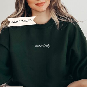 Puede incluir: Sudadera verde bosque con la frase bordada "most ardently". La parte superior tiene cuello redondo y mangas largas. La palabra "EMBROIDERED!" est&aacute; impresa en una pancarta blanca. La palabra "Forest" est&aacute; impresa en una pancarta blanca.