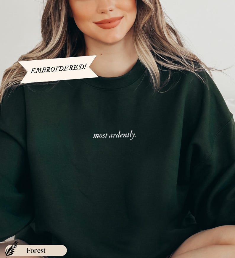 Puede incluir: Sudadera verde bosque con la frase bordada "most ardently". La parte superior tiene cuello redondo y mangas largas. La palabra "EMBROIDERED!" est&aacute; impresa en una pancarta blanca. La palabra "Forest" est&aacute; impresa en una pancarta blanca.