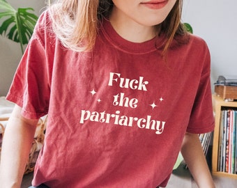 May include: A maroon-colored t-shirt with the text "Fuck the patriarchy" in white, along with two star graphics. The shirt is a casual style, likely made of cotton or a similar fabric.