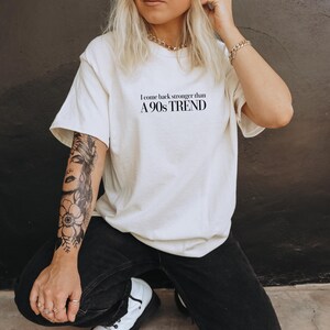 Op de afbeelding: Een wit T-shirt met zwarte tekst die "Ik kom sterker terug dan een trend uit de jaren 90" luidt. De persoon die het shirt draagt heeft een zwart-witte tatoeage op zijn arm.