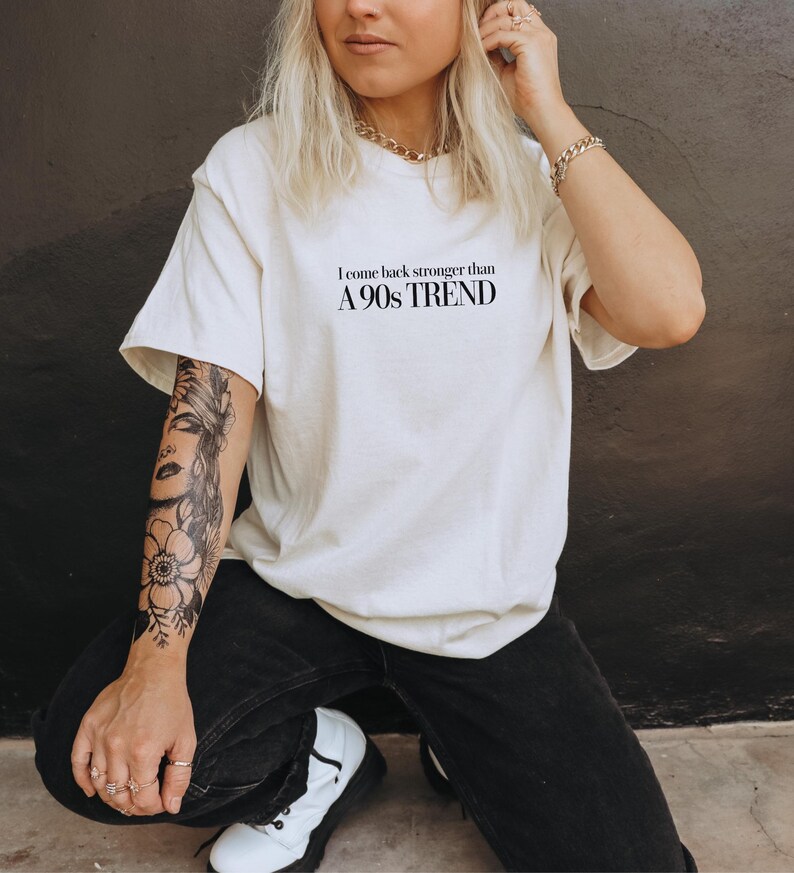 Op de afbeelding: Een wit T-shirt met zwarte tekst die "Ik kom sterker terug dan een trend uit de jaren 90" luidt. De persoon die het shirt draagt heeft een zwart-witte tatoeage op zijn arm.