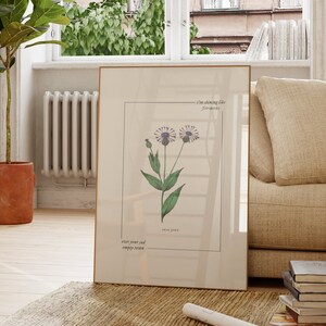 May include: A framed botanical print with a quote that reads "I am choosing life, forever." The print features a drawing of a purple flower with green leaves and stems. The quote is written in a simple font and is centered below the flower.