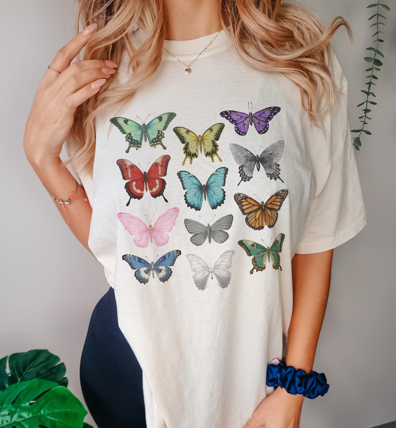 May include: Cream-colored t-shirt featuring a colorful butterfly design. The butterflies are in various colors, including green, yellow, red, blue, pink, and orange. The butterflies are arranged in a grid pattern across the front of the shirt.