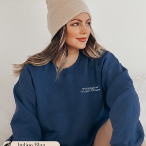 May include: An indigo blue crewneck sweatshirt featuring the white text Pathological People Pleaser printed on the upper left chest. The garment has long sleeves and a ribbed neckline. A beige knit beanie is worn with the indigo blue sweatshirt.