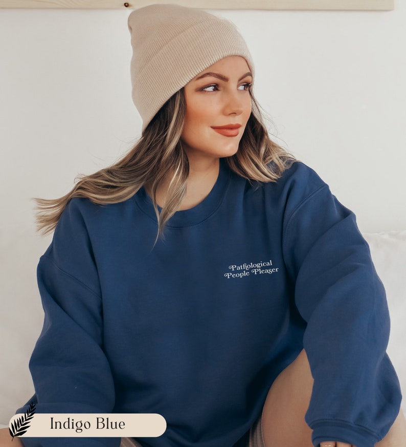 May include: An indigo blue crewneck sweatshirt featuring the white text Pathological People Pleaser printed on the upper left chest. The garment has long sleeves and a ribbed neckline. A beige knit beanie is worn with the indigo blue sweatshirt.