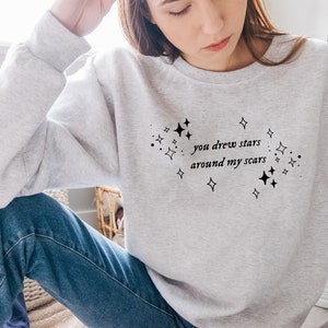 May include: A gray sweatshirt with the text "you drew stars around my scars" printed on the front. The text is surrounded by small stars.