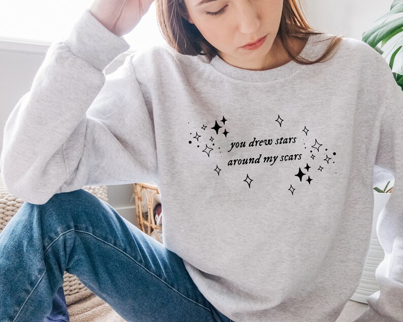 May include: A gray sweatshirt with the text "you drew stars around my scars" printed on the front. The text is surrounded by small stars.