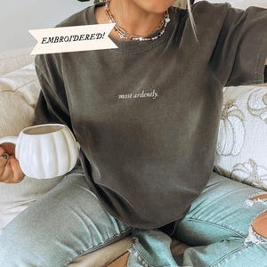 Op de afbeelding: Een oversized donkergrijs T-shirt met de tekst "most ardently." geborduurd op de voorkant. Het shirt wordt gedragen door een persoon die op een bank zit.