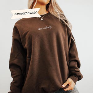 Puede incluir: Una sudadera bordada de color marr&oacute;n oscuro con las palabras "most ardently" en escritura blanca. La sudadera tiene mangas largas y cuello redondo. La palabra "EMBROIDERED!" est&aacute; en una pancarta encima de la sudadera.