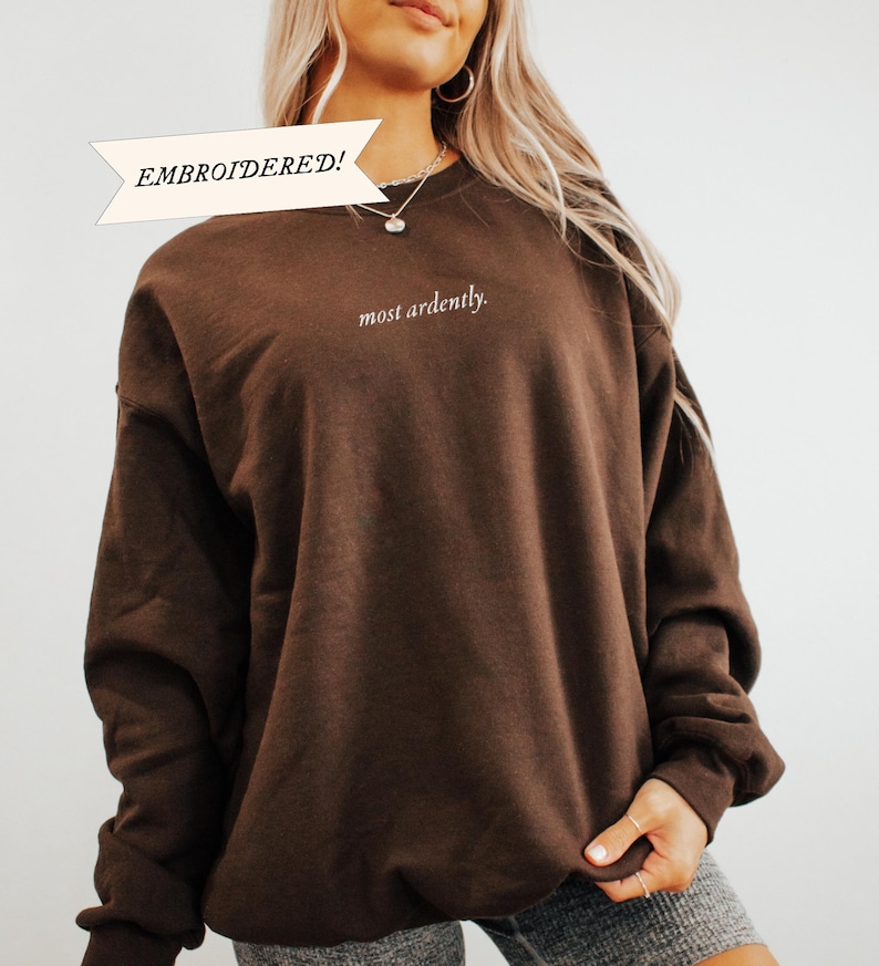 Puede incluir: Una sudadera bordada de color marr&oacute;n oscuro con las palabras "most ardently" en escritura blanca. La sudadera tiene mangas largas y cuello redondo. La palabra "EMBROIDERED!" est&aacute; en una pancarta encima de la sudadera.