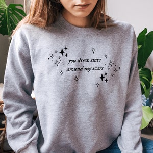 May include: Gray sweatshirt with a black graphic print that says "you drew stars around my scars" with small stars surrounding the text.