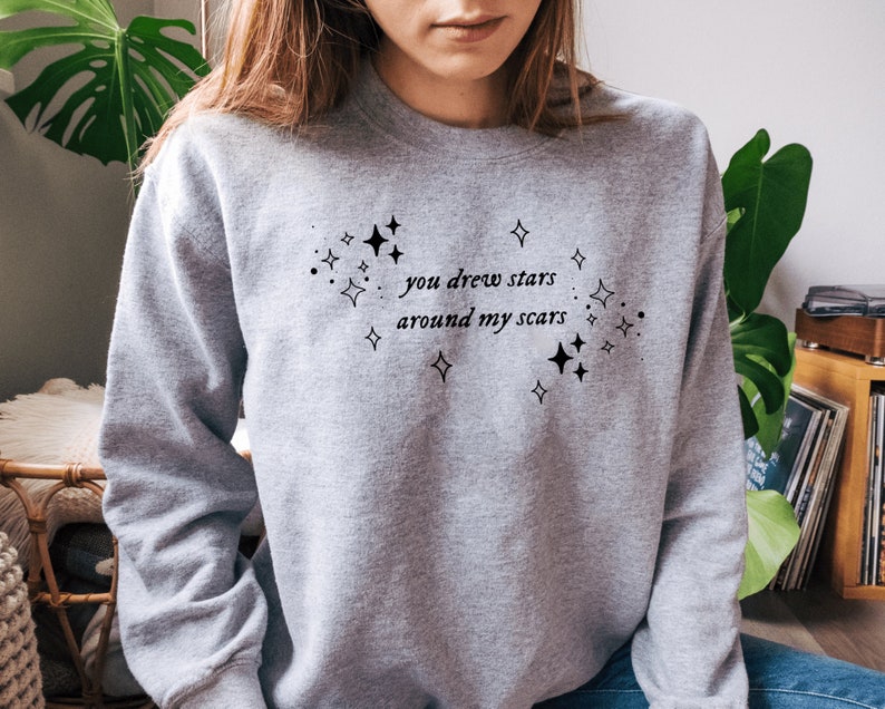 May include: Gray sweatshirt with a black graphic print that says "you drew stars around my scars" with small stars surrounding the text.