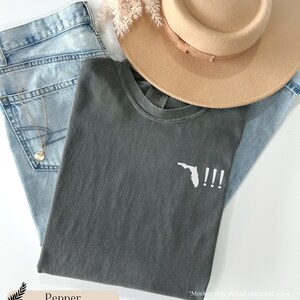 Peut inclure: Un t-shirt gris fonc&eacute; avec un contour blanc de l'&eacute;tat de Floride et trois points d'exclamation. Le t-shirt est pli&eacute; et pos&eacute; sur un jean bleu clair et un chapeau beige.