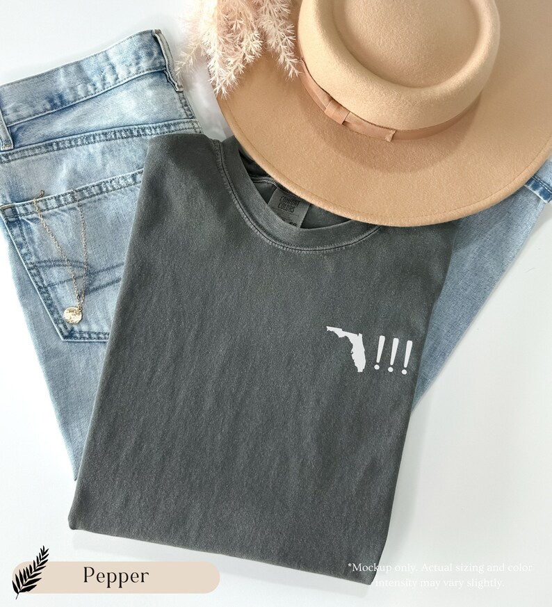 Peut inclure: Un t-shirt gris fonc&eacute; avec un contour blanc de l'&eacute;tat de Floride et trois points d'exclamation. Le t-shirt est pli&eacute; et pos&eacute; sur un jean bleu clair et un chapeau beige.