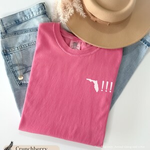 Peut inclure: Un t-shirt rose avec un contour blanc de l'&eacute;tat de Floride et trois points d'exclamation. Le t-shirt est pos&eacute; &agrave; plat sur un fond en jean bleu clair.