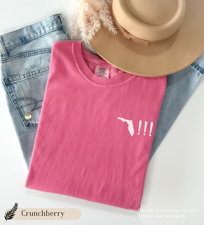 Peut inclure: Un t-shirt rose avec un contour blanc de l'&eacute;tat de Floride et trois points d'exclamation. Le t-shirt est pos&eacute; &agrave; plat sur un fond en jean bleu clair.