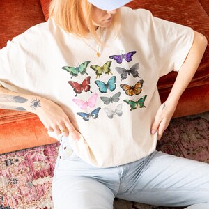 May include: Cream-colored t-shirt featuring a colorful butterfly graphic. The butterflies are in various colors, including green, blue, red, and purple. The shirt is paired with light blue jeans and a light blue baseball cap. The overall aesthetic is casual and artistic.
