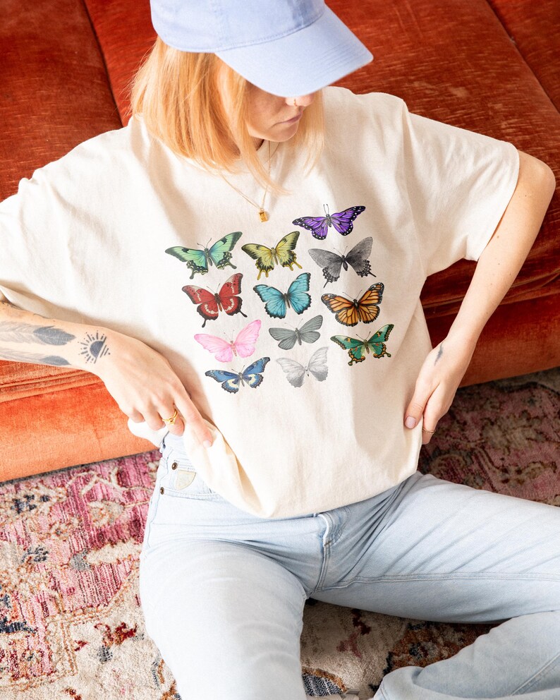May include: Cream-colored t-shirt featuring a colorful butterfly graphic. The butterflies are in various colors, including green, blue, red, and purple. The shirt is paired with light blue jeans and a light blue baseball cap. The overall aesthetic is casual and artistic.