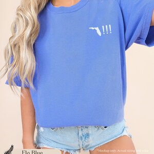Peut inclure: Un t-shirt bleu clair avec un contour blanc de l'&eacute;tat de Floride et trois points d'exclamation. Le texte "Flo Blue" est visible en bas de l'image.