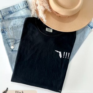 Peut inclure: T-shirt noir avec un contour blanc de l'&eacute;tat de Floride et trois points d'exclamation. Le texte "COMFORT COLORS" est visible sur l'&eacute;tiquette du t-shirt.