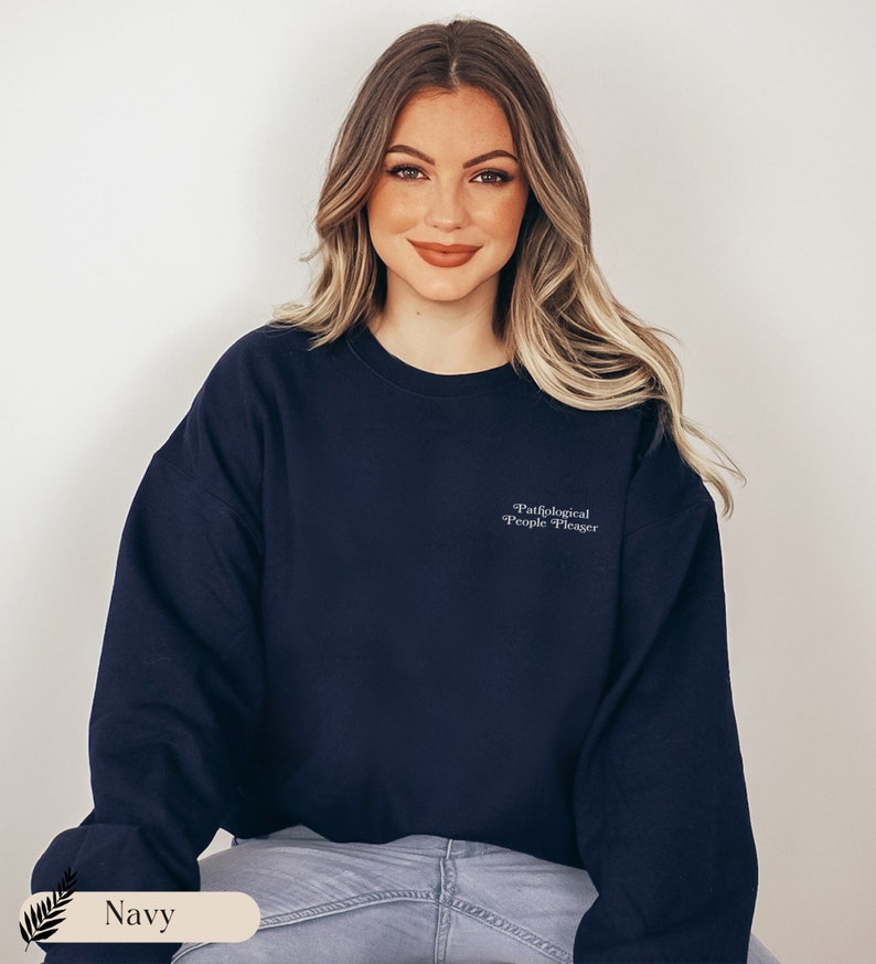 May include: A navy blue crewneck sweatshirt featuring the white embroidered text "Pathological People Pleaser" on the upper left chest. The garment has long sleeves, a ribbed neckline, and a classic fit, providing a simple and clean aesthetic.
