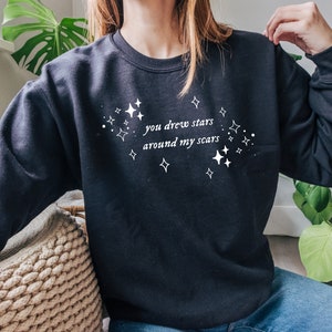May include: Black sweatshirt with white text that reads "you drew stars around my scars" and a design of white stars.