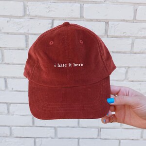 May include: A rust-colored corduroy baseball cap with the phrase "i hate it here" embroidered in white. The cap has a curved brim and a rounded crown. The hat is held by a hand with blue nail polish.