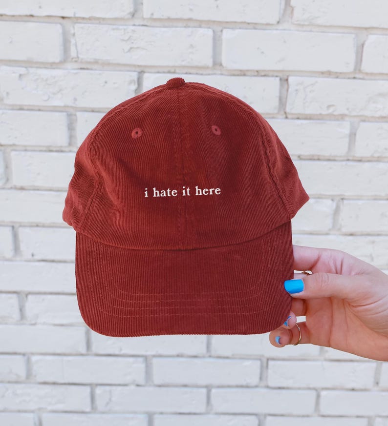 May include: A rust-colored corduroy baseball cap with the phrase "i hate it here" embroidered in white. The cap has a curved brim and a rounded crown. The hat is held by a hand with blue nail polish.