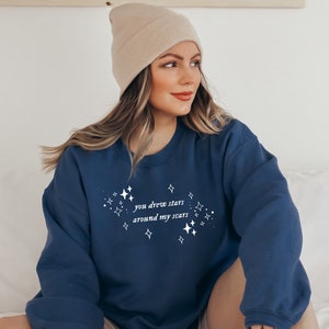 May include: A navy blue sweatshirt with the text "you drew stars around my scars" in white lettering and a design of white stars around the text.
