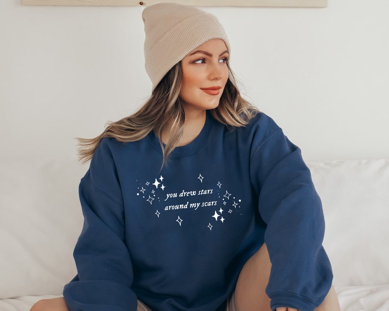 May include: A navy blue sweatshirt with the text "you drew stars around my scars" in white lettering and a design of white stars around the text.