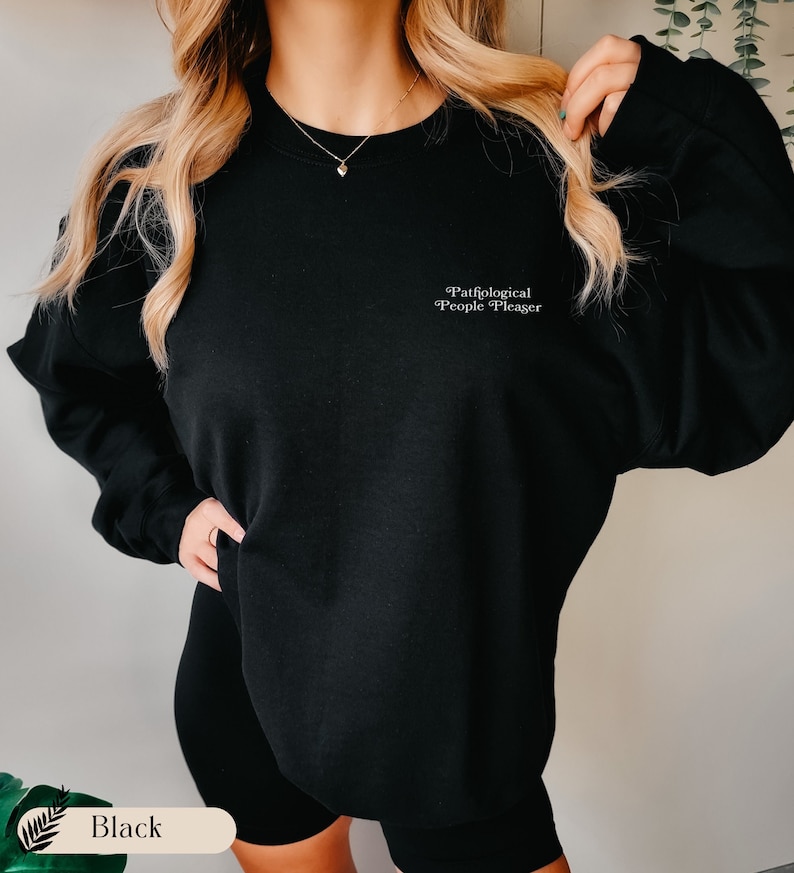 May include: A black crewneck sweatshirt featuring the white text Pathological People Pleaser printed on the upper left chest area. The garment has long sleeves and a standard fit, providing a simple and clean look for casual daily wear.