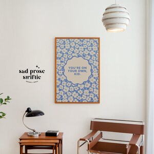 May include: A framed print with a blue and white floral pattern and the text "You're on your own, kid." hanging on a white wall. A small plant, a lamp, and a chair are also in the image.