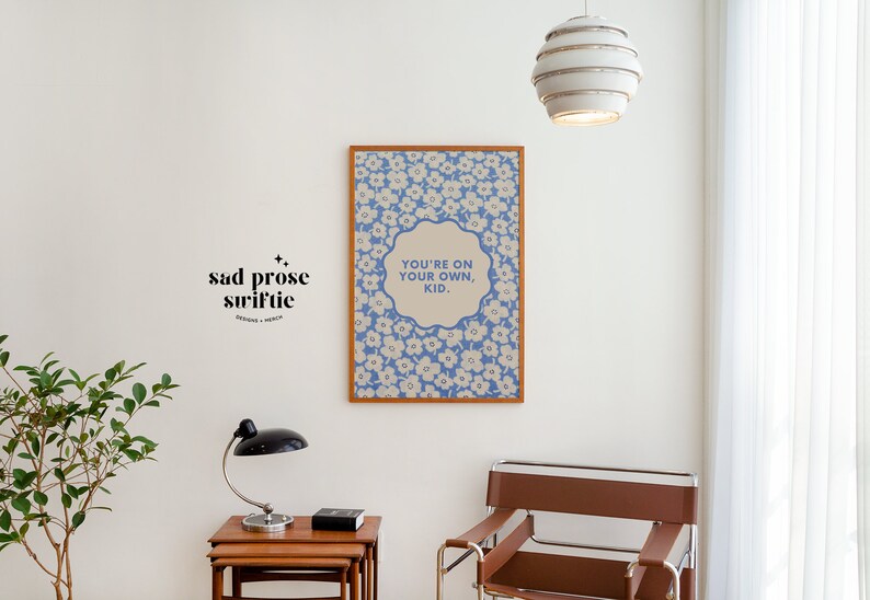 May include: A framed print with a blue and white floral pattern and the text "You're on your own, kid." hanging on a white wall. A small plant, a lamp, and a chair are also in the image.