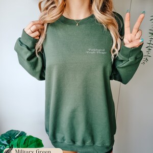 May include: A military green crewneck sweatshirt featuring long sleeves and ribbed cuffs. The text 'Pathological People Pleaser' is printed in a small, white, serif font on the upper left chest area of the garment.