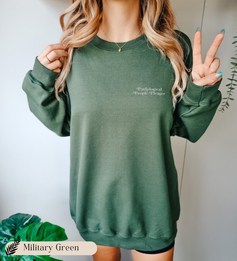 May include: A military green crewneck sweatshirt featuring long sleeves and ribbed cuffs. The text 'Pathological People Pleaser' is printed in a small, white, serif font on the upper left chest area of the garment.