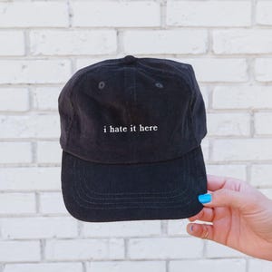May include: Black corduroy baseball cap with the phrase "i hate it here" embroidered in white. The cap has a curved brim and a structured crown. The hat is held up against a white brick wall.