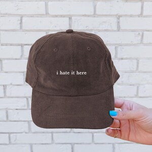 May include: A brown corduroy baseball cap with the phrase "i hate it here" embroidered in white. The cap has a curved brim and a button on top. The hat is a solid brown color.