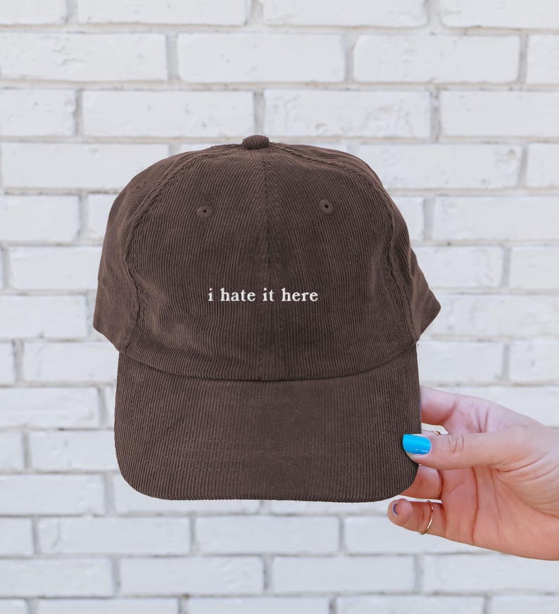 May include: A brown corduroy baseball cap with the phrase "i hate it here" embroidered in white. The cap has a curved brim and a button on top. The hat is a solid brown color.