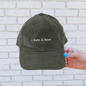 May include: Olive green corduroy baseball cap with the embroidered text "i hate it here" in white. The cap has a curved brim and a structured crown. The hat is a classic six-panel design.