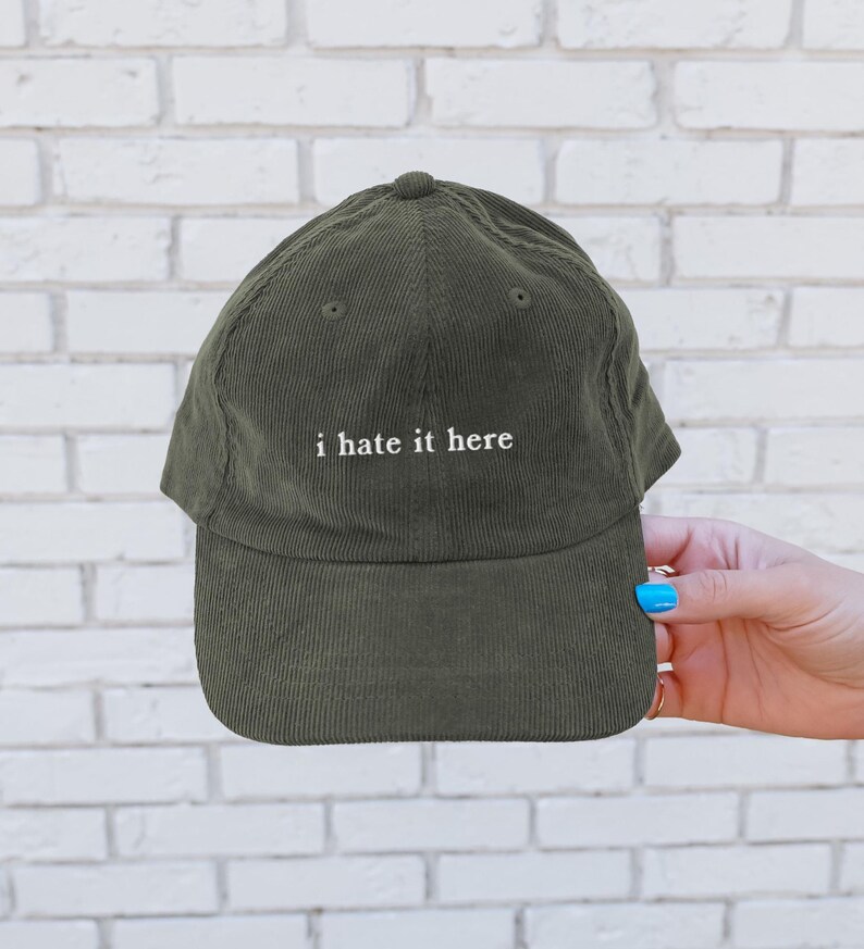 May include: Olive green corduroy baseball cap with the embroidered text "i hate it here" in white. The cap has a curved brim and a structured crown. The hat is a classic six-panel design.