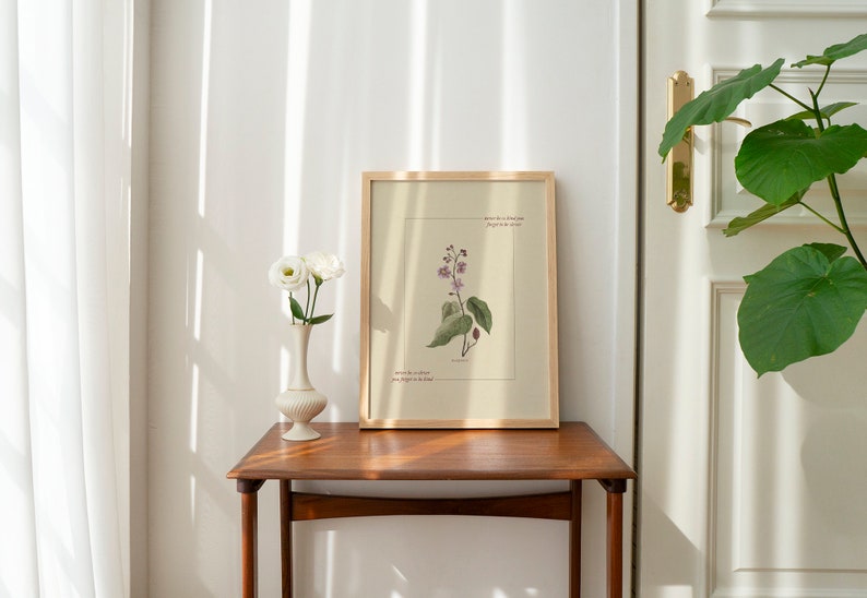 May include: A framed botanical illustration of a pink flower with green leaves. The frame is made of wood and has a light brown finish. The illustration is on a cream-colored background. The text on the illustration reads "Where do we find that old magic? In the details."