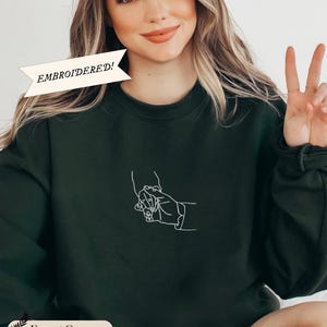 Może przedstawiać: Zielony sweter z nadrukiem w kolorze białym przedstawiającym dwie trzymające się za ręce dłonie. Tekst "EMBROIDERED!" znajduje się na białym banerze u góry obrazu.