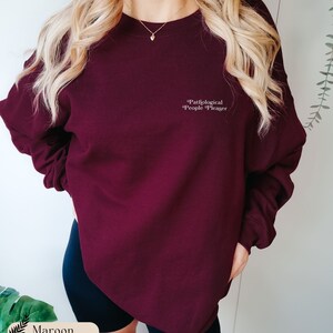 May include: A maroon crewneck sweatshirt featuring the white text Pathological People Pleaser printed on the upper left chest. The garment has long sleeves with ribbed cuffs and a ribbed neckline, providing a simple and clean look for casual wear.