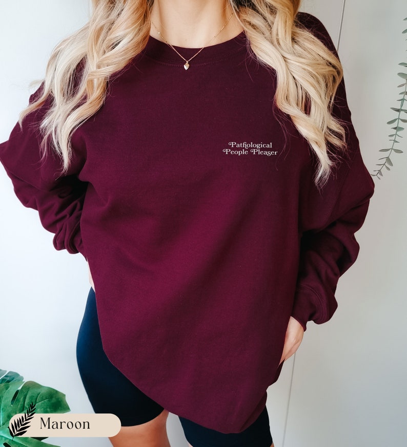 May include: A maroon crewneck sweatshirt featuring the white text Pathological People Pleaser printed on the upper left chest. The garment has long sleeves with ribbed cuffs and a ribbed neckline, providing a simple and clean look for casual wear.