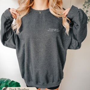 May include: A dark heather gray crewneck sweatshirt featuring the text Pathological People Pleaser embroidered in white on the upper left chest. The garment has long sleeves, a ribbed neckline, and a relaxed fit with a soft, textured fabric finish.