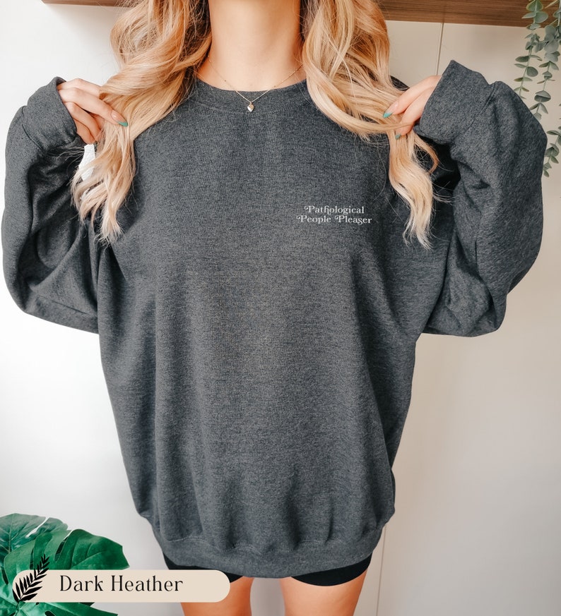 May include: A dark heather gray crewneck sweatshirt featuring the text Pathological People Pleaser embroidered in white on the upper left chest. The garment has long sleeves, a ribbed neckline, and a relaxed fit with a soft, textured fabric finish.