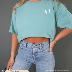 Peut inclure: T-shirt bleu turquoise avec un contour blanc de l'&eacute;tat de Floride et trois points d'exclamation. Le t-shirt est port&eacute; par une personne portant un jean bleu.