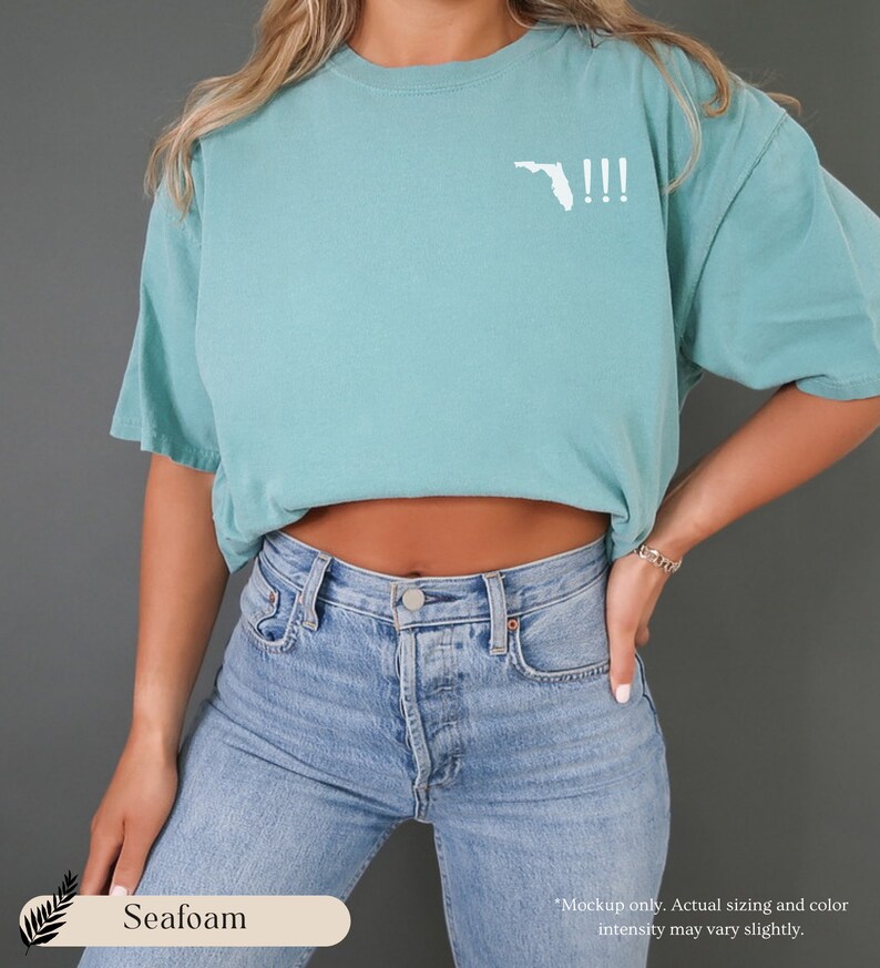 Peut inclure: T-shirt bleu turquoise avec un contour blanc de l'&eacute;tat de Floride et trois points d'exclamation. Le t-shirt est port&eacute; par une personne portant un jean bleu.