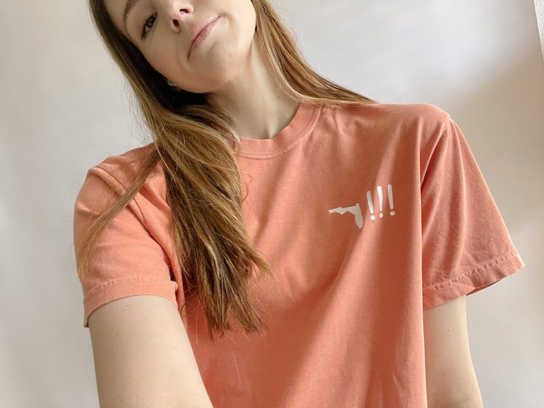 Peut inclure: Un t-shirt de couleur corail avec un contour blanc de l'&eacute;tat de Floride et trois points d'exclamation.