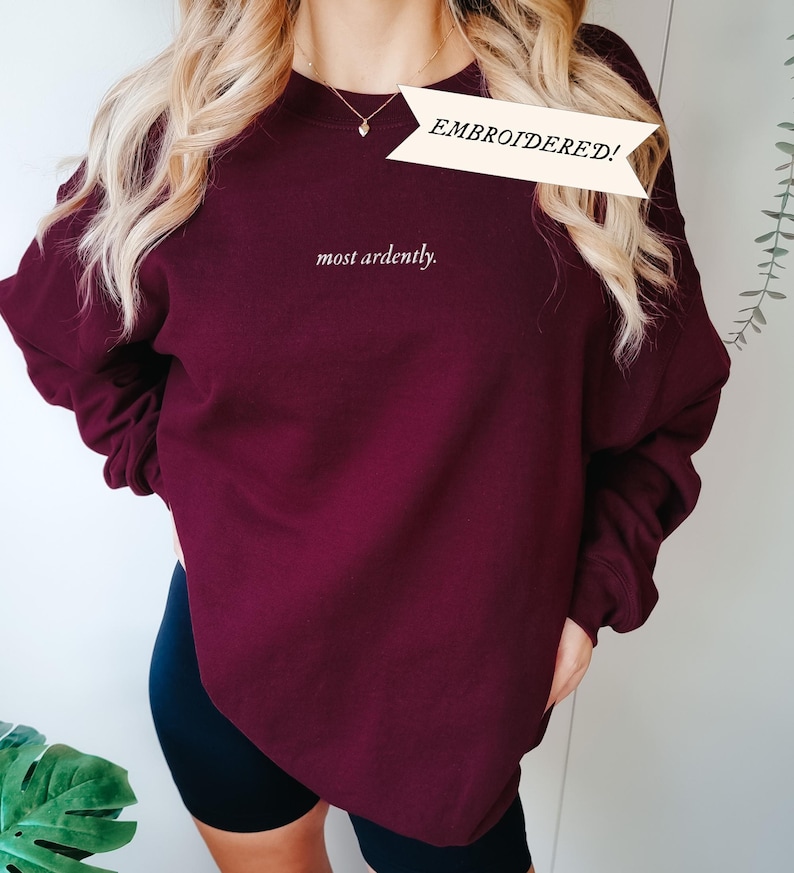 Puede incluir: Sudadera burdeos con el texto bordado "most ardently". Una pancarta sobre el texto dice "&iexcl;BORDADO!". La sudadera es de color liso y tiene mangas largas. La modelo lleva un pantal&oacute;n corto negro.